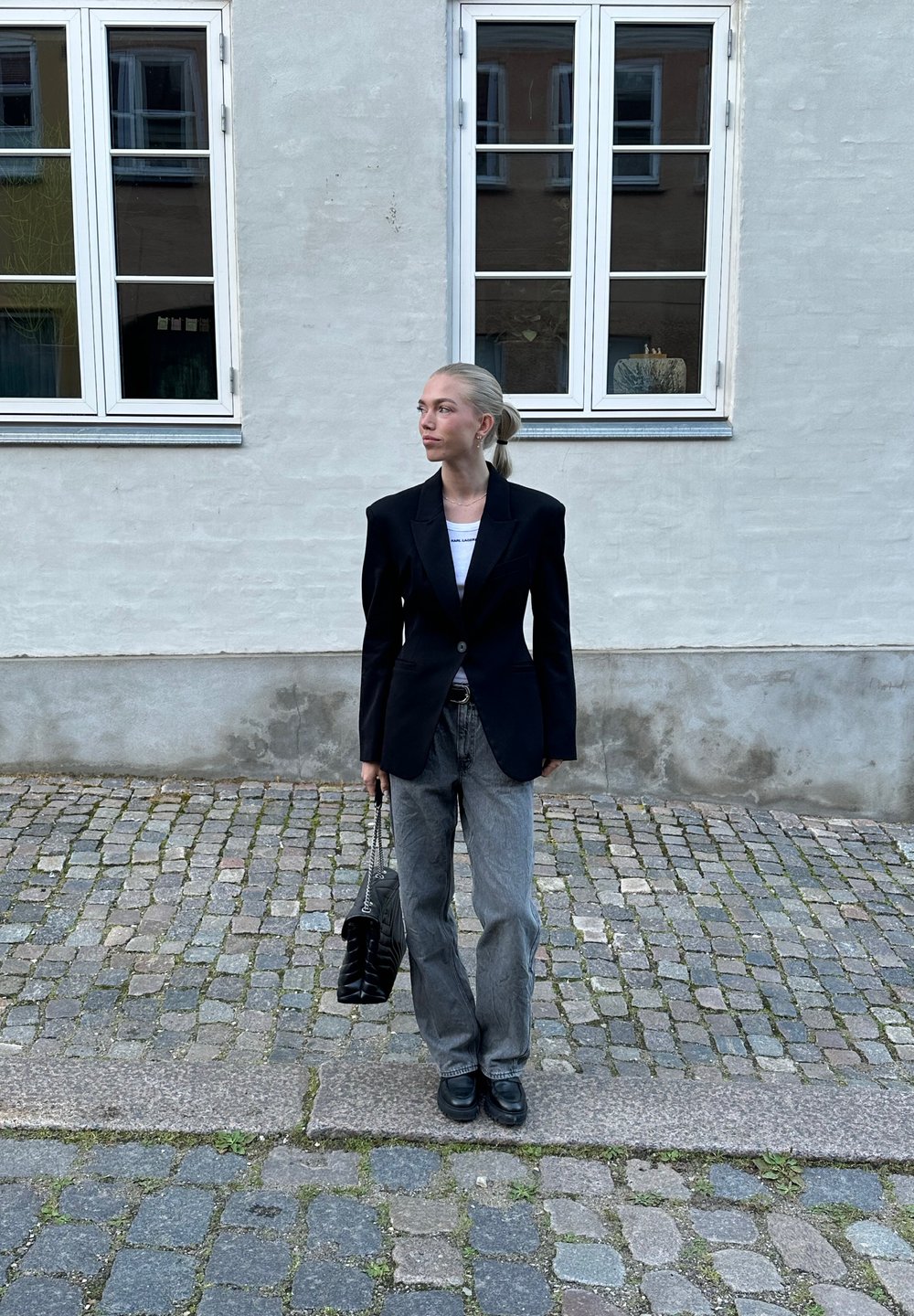 Μαύρο εφαρμοστό σακάκι, λευκή μπλούζα και γκρι φαρδιά τζιν παντελόνια. Αξεσουάρ με μια μαύρη τσάντα με καπιτονέ σχέδιο και μαύρα παπούτσια. Πλακόστρωτο δάπεδο.