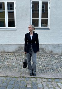 Μαύρο εφαρμοστό σακάκι, λευκή μπλούζα και γκρι φαρδιά τζιν παντελόνια. Αξεσουάρ με μια μαύρη τσάντα με καπιτονέ σχέδιο και μαύρα παπούτσια. Πλακόστρωτο δάπεδο.