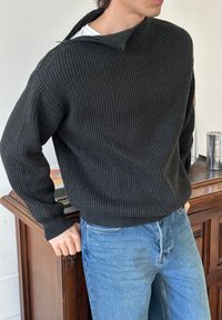 Mann som har på seg en mørk ribbestrikket genser med glidelåshals, stukket inn i lyseblå jeans, står nær et trebords skap med metallisk dekor.