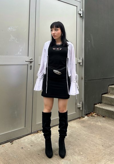 Vestido negro con un detalle recortado, superpuesto con un cárdigan de manga de campana en gris claro. Accesorios incluyen un cinturón plateado y botas negras hasta la rodilla.