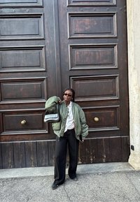 Il modello indossa una giacca bomber verde, una camicia bianca e pantaloni neri. Tiene una borsa nera. Lo sfondo presenta grandi porte in legno con accessori dorati.