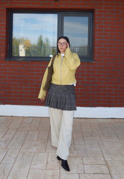 Mujer con chaqueta amarilla, falda a cuadros sobre pantalones beige, botas negras, gafas y bolso marrón al hombro, de pie sobre un suelo de baldosas.