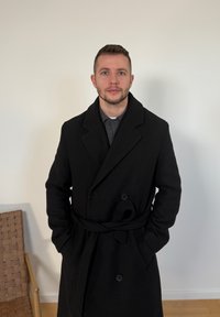 Άνδρας με κοντά μαλλιά και γένια, που φοράει μαύρο παλτό με ζώνη, στέκεται με τα χέρια στις τσέπες μπροστά σε έναν απλό λευκό τοίχο.