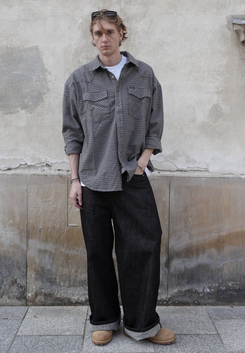 Jeune homme avec des lunettes de soleil sur la tête, portant une chemise grise à carreaux ample, un pantalon noir large et des bottes beige, debout contre un mur en béton usé.