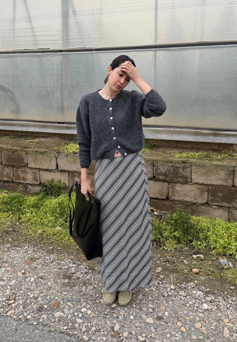 Woman with dark hair in gray buttoned sweater and striped skirt, holding black bag, standing on gravel with concrete wall background.