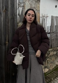 Γυναίκα που φοράει σκούρο μπουφάν και γκρι πλισέ φούστα, κρατώντας μια λευκή υφαντή τσάντα, στέκεται μπροστά σε φόντο πέτρινης και ξύλινης περίφραξης.