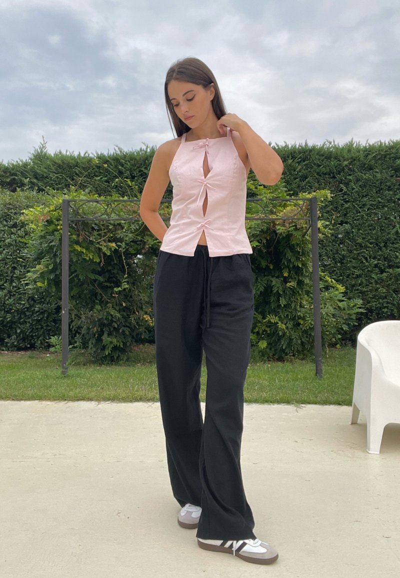 Rosa top a fascia con ritagli a forma di chiave, abbinato a pantaloni neri a gamba larga. La modella si trova su un patio con sfondo verde.