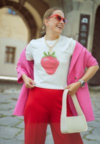Balta marškinėliai su dideliu raudonu braškių grafiku, rožinė džinsinė švarkas, raudoni plačiakelniai kelnės, smėlio spalvos karoliukų kaklo papuošalas ir baltas rankinukas. Raudoni saulės akiniai.