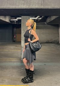 Top negru fără mâneci cu știfturi, fustă cu dungi negre și albe, geantă de umăr neagră și cizme masive negre cu catarame, amplasate într-un garaj auto.