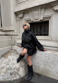 Schwarze Lederjacke in Übergröße, schwarzes Minikleid, kniehohe schwarze Stiefel. Glatte Textur, minimalistisches Design, inszeniert vor einer Granitwand.