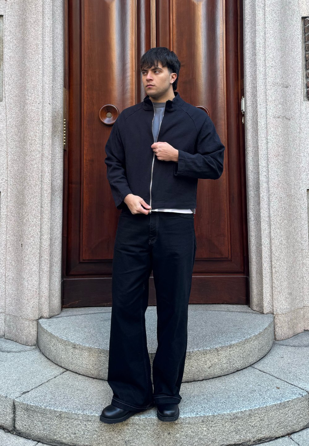 Jeune homme en veste noire zippée, pantalon large noir et bottes, debout sur des marches en pierre devant une grande porte en bois.