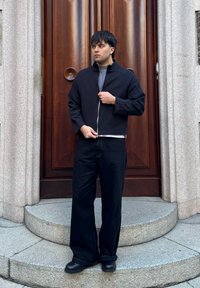 Jeune homme en veste noire zippée, pantalon large noir et bottes, debout sur des marches en pierre devant une grande porte en bois.