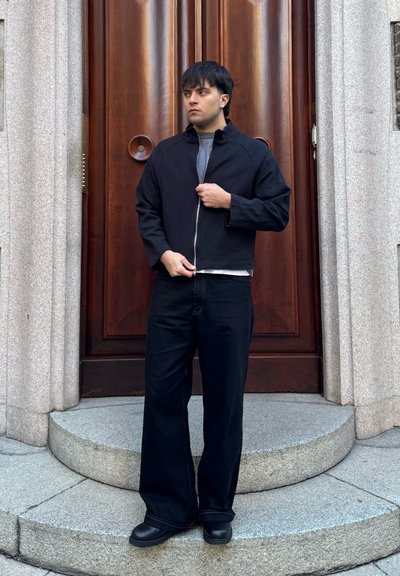 Joven con chaqueta negra con cremallera, pantalones negros anchos y botas, de pie sobre escalones de piedra frente a una gran puerta de madera.