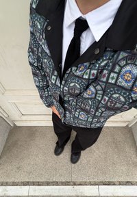 Veste colorée avec un motif en vitrail vert, bleu et orange, dotée d'un col noir. Portée sur une chemise blanche et une cravate noire.