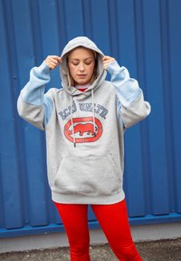 Γκρίζα ζακέτα με μπλε μανίκια, διαθέτει το λογότυπο Ecko United σε κόκκινο και μαύρο χρώμα. Έχει μπροστινή τσέπη και κουκούλα με κορδόνι. Φοριέται με κόκκινα κολάν.