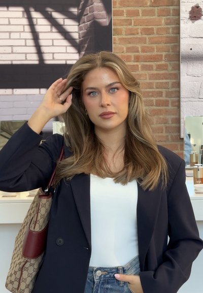 Mujer joven con cabello rubio largo, ojos azules, vestida con un blazer negro y una blusa blanca, sosteniendo un bolso con estampado, de pie en el interior.