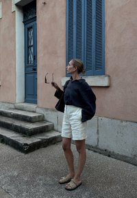 Tengerész csíkos, túlméretezett blúz, fehér vászon rövidnadrág, bézs szandál. Fekete táska és napszemüveg a kézben. Texturált rózsaszín fal a háttérben.