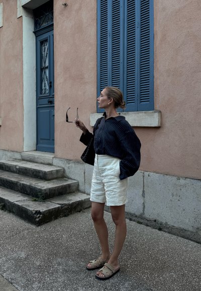 Blusa de rayas marinas oversized, pantalones cortos de lino blancos, sandalias beiges. Bolsa negra y gafas de sol sujetas con la mano. Pared texturizada rosa de fondo.