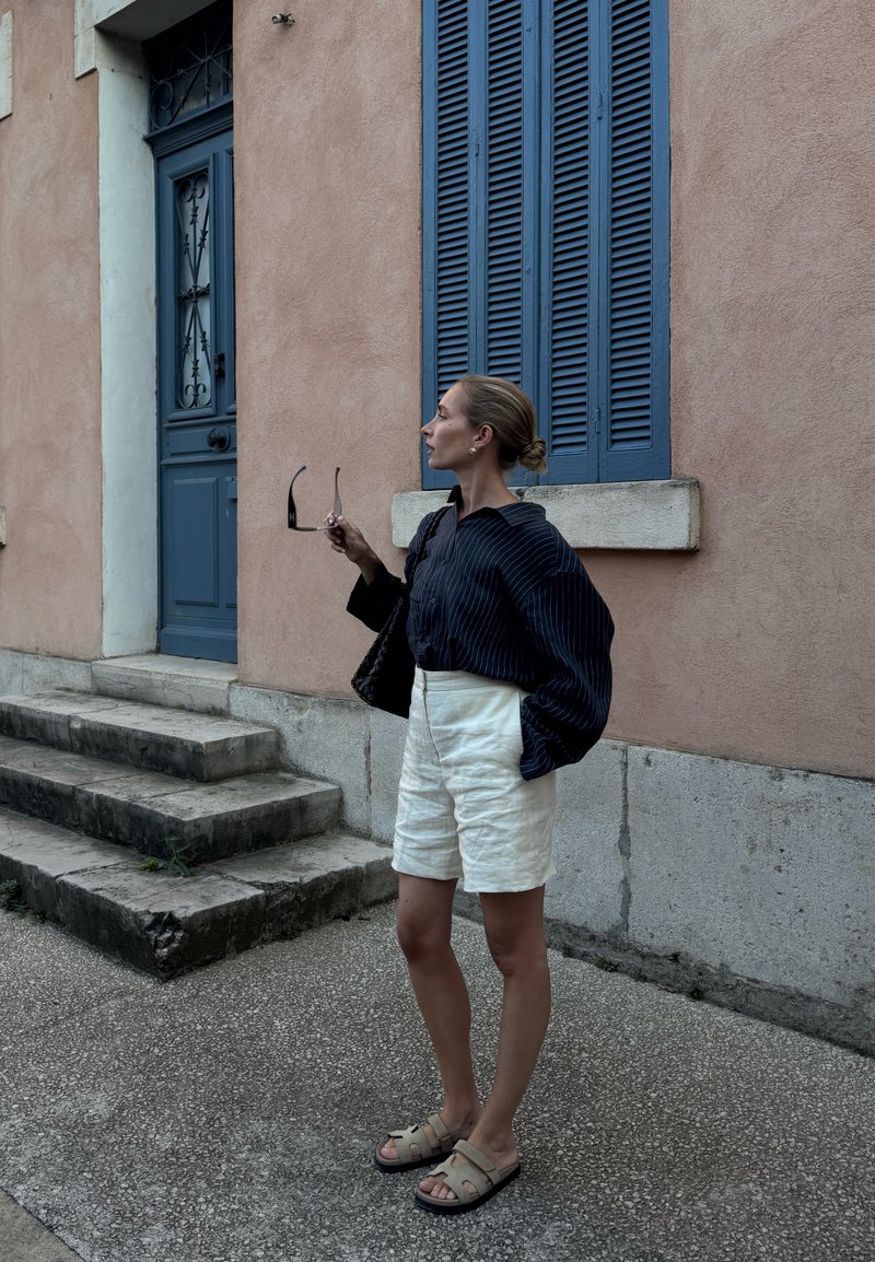 Blusa oversize a righe navy, pantaloni corti di lino bianchi, sandali beige. Borsa nera e occhiali da sole tenuti in mano. Parete rosa testurizzata sullo sfondo.