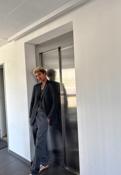 Persona apoyada contra una pared cerca de un ascensor plateado. Lleva un blazer de rayas, una camisa gris, pantalones amplios grises y zapatillas claras.