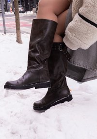 Άτομο που φοράει μαύρες δερμάτινες μπότες μέχρι το γόνατο και ένα ανοιχτό μπεζ χνουδωτό μπουφάν, στέκεται στο χιόνι κοντά σε μια γωνία δρόμου.