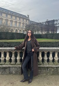 Γυναίκα με σκούρα μαλλιά, γυαλιά, καφέ μακρύ παλτό και μαύρες μπότες που ακουμπά σε πέτρινο μπαλκόνι μπροστά σε φράκτες και ιστορικό κτίριο κάτω από συννεφιασμένο ουρανό.