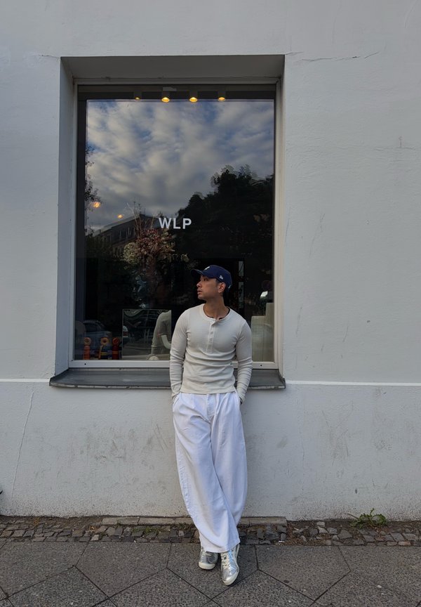 Homme portant une chemise blanche, un pantalon blanc large et une casquette marine, appuyé contre un mur blanc sous une fenêtre avec les lettres WLP.