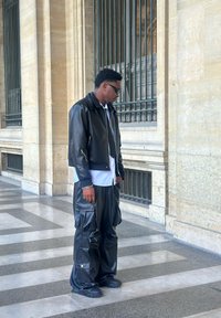 Black leather cropped bomber jacket with silver zippers, paired with loose black baggy pants featuring side pockets. White shirt underneath.