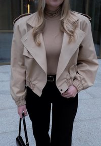 Veste bomber beige avec un large col, des poignets élastiqués et une coupe décontractée. Associée à un haut beige ajusté et un pantalon noir.