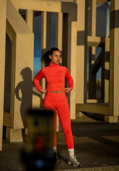 Conjunto deportivo de color naranja brillante, camiseta ajustada de manga larga, leggings a juego, textura suave, con cuello con cremallera y acentos de logo. Sombra en el suelo.