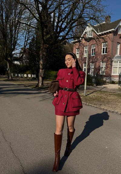 Mujer caminando por una calle vistiendo un vestido rojo con botones y botas marrones hasta la rodilla, sosteniendo un bolso marrón, con árboles y edificios de ladrillo de fondo.