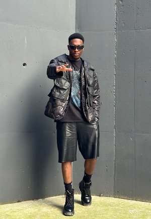 Chaqueta negra acolchada con múltiples bolsillos, camiseta gráfica negra, pantalones cortos de cuero negros y botas de combate negras. Con un fondo de pared gris.