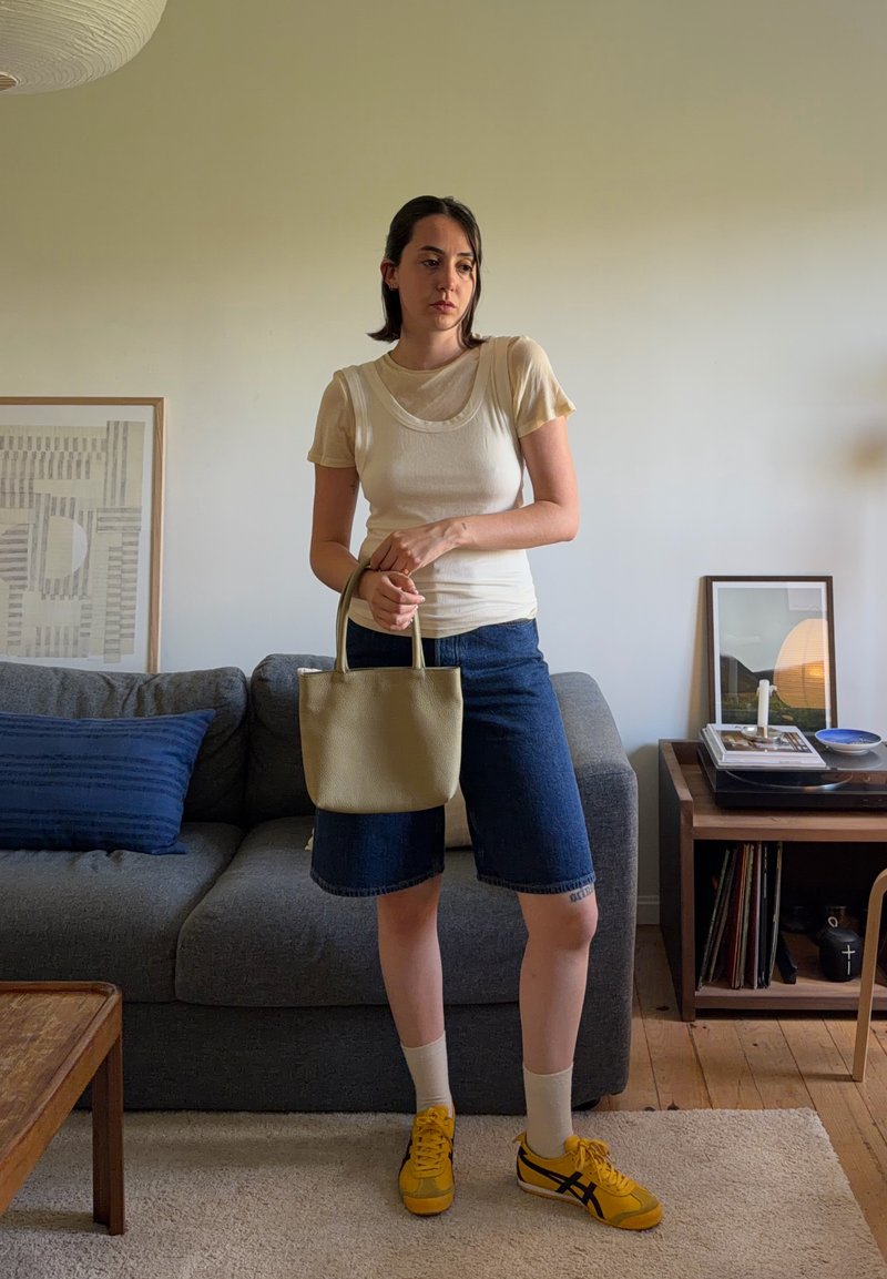 Jeune femme en tenue décontractée tenant un sac à main beige, debout dans un salon avec un canapé gris et des meubles en bois.