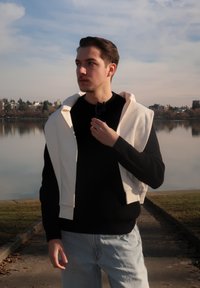 Νέος άνδρας με μαύρο πουλόβερ και ανοιχτόχρωμο τζιν κρατά γυαλιά ηλίου, με λευκό μπουφάν περασμένο στους ώμους, στέκεται δίπλα σε ήρεμη λίμνη κάτω από συννεφιασμένο ουρανό.