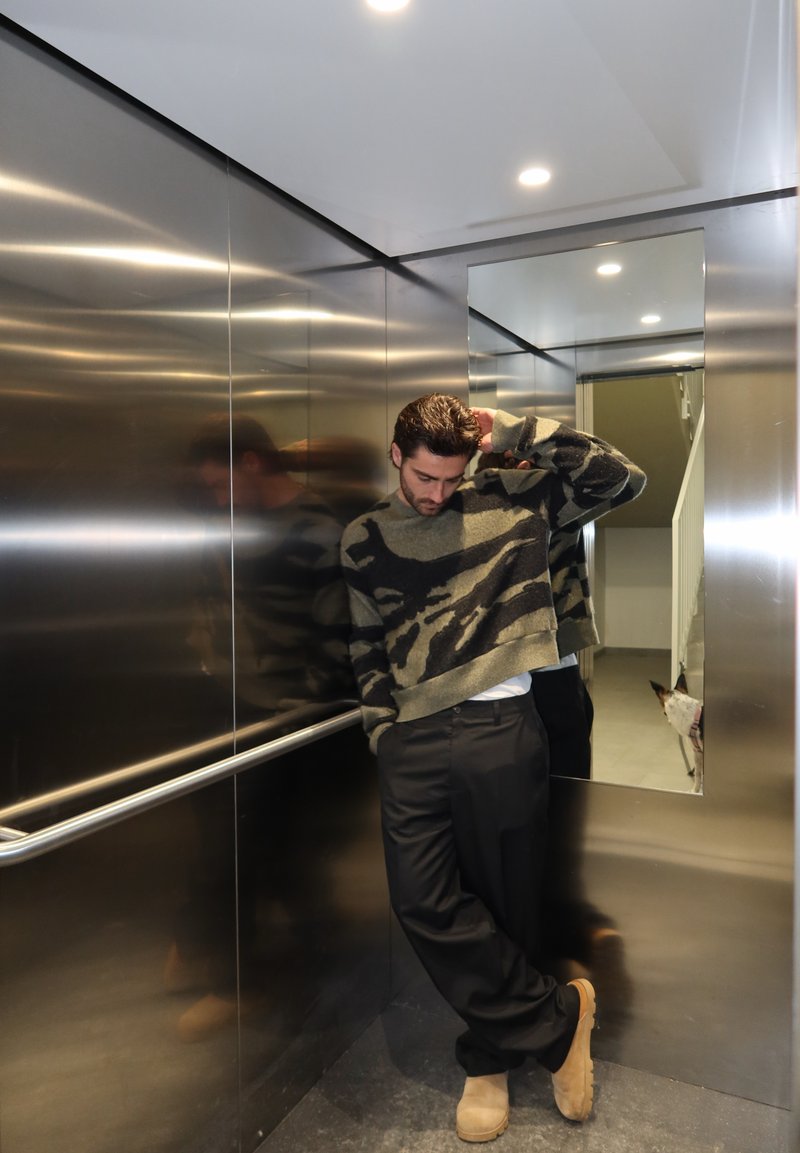 Άνδρας με καμουφλαρισμένο πουλόβερ στηρίζεται στον τοίχο του ανελκυστήρα με το κεφάλι σκυμμένο· ένας σκύλος φαίνεται μερικώς στον καθρέφτη κοντά στην πόρτα του ανελκυστήρα.