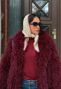 Femme portant un manteau en fausse fourrure bordeaux surdimensionné, un pull en maille marron foncé, un foulard en crochet crème, des lunettes de soleil sombres, debout près d'une porte marron.