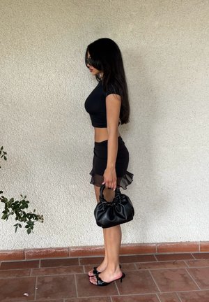 Schwarzes Crop-Top, transparente schwarze Minirock mit Rüschensaum, schwarze schlanke Handtasche und schwarze Riemchensandalen mit Schleifendetail.