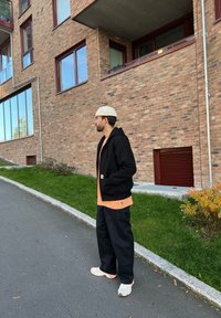 Black zip-up hoodie over an orange t-shirt, teamed with loose black trousers and white trainers. The background showcases a brick wall and windows.