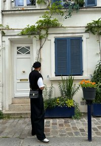 Μαύρη τσάντα ώμου, λευκό μπλουζάκι και μαύρο παντελόνι φορεμένα από ένα άτομο κοντά σε έναν λευκό τοίχο, σκούρες μπλε περσίδες και πολύχρωμες γλάστρες φυτών.