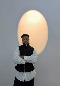 Fekete kötött mellény fehér ing felett, karok összefonva. A háttérben nagy ovális fény látható egy lágy szürke fal előtt.