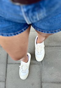 Zapatillas blancas con acentos beiges, con un diseño de cordones, situadas sobre un pavimento gris junto a unos pantalones cortos de mezclilla. Textura suave.