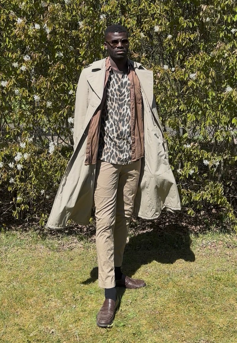 Homme portant des lunettes de soleil, trench beige sur une chemise à imprimé animalier et pantalon beige, debout sur l'herbe avec des buissons verts feuillus derrière.
