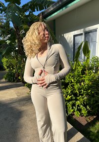 Beige zip-up long-sleeved top with fitted sleeves, paired with matching fitted trousers. Smooth, stretchy fabric in natural light. Green foliage in the background.