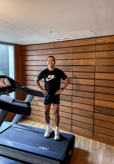 Hombre con ropa de correr negra de pie en una cinta de correr en un gimnasio con paredes de paneles de madera y una gran ventana.