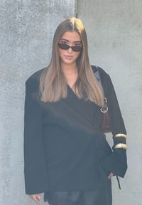 Melna ārējā apģērba oversized dizains ar zelta akcentiem uz piedurknēm, kombinēts ar tumšu satīna svārku un mazām melnām saulesbrillēm.