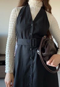 Rochie neagră tip halter cu nasturi pe partea din față și o curea din material textil. Helancă albă texturată și o geantă maro închis cu mâner curbat.