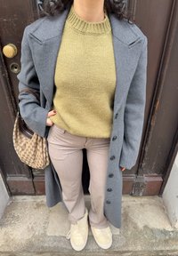 Long manteau gris par-dessus un pull en tricot vert, pantalon beige et chaussures jaunes. Accentué par un sac à main marron à motifs.