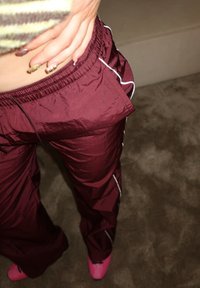 Pantalon de survêtement bordeaux à la texture brillante, avec des bandes blanches sur les côtés et une taille élastique, associé à des chaussures roses.