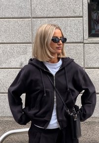 Felpa nera con zip e vestibilità rilassata, t-shirt bianca sotto e occhiali da sole neri. Una piccola borsa a tracolla nera pende su una spalla.
