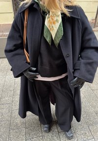 Melna vilnas mēteli, tumši džemperi, balta krekls un platās bikses; zaļš un krēmkrāsas rakstains šalle; melnas cimdi; brūna plecu soma.